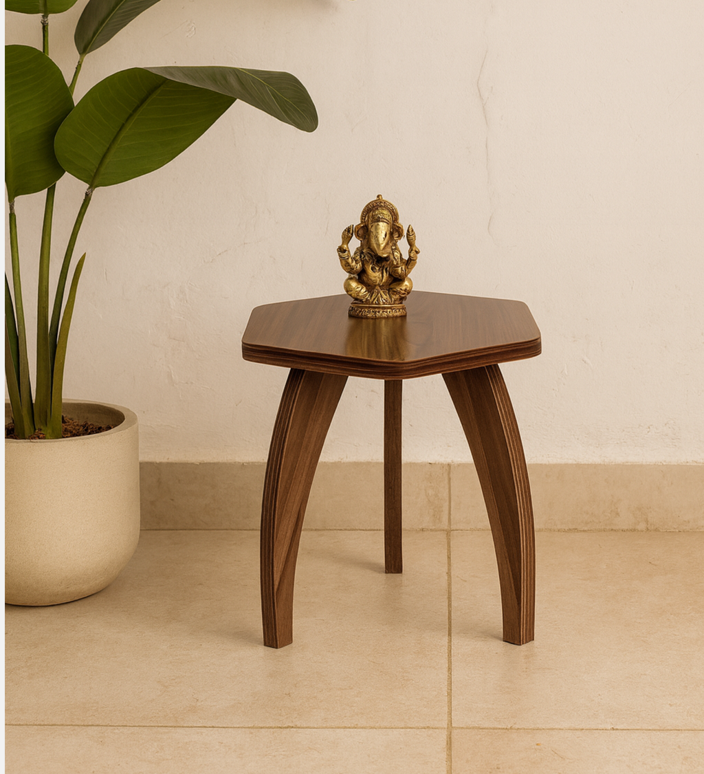A small hexagonal wooden side table with three curved legs, styled in a home setting next to a potted plant with a brass Ganesha statue on top, set against a beige wall and tiled floor.